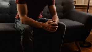 Zu sehen ist ein Mann, von den Schultern abwärts, der auf einer Couch sitzt und die Hände zum Beten gefaltet hat.
