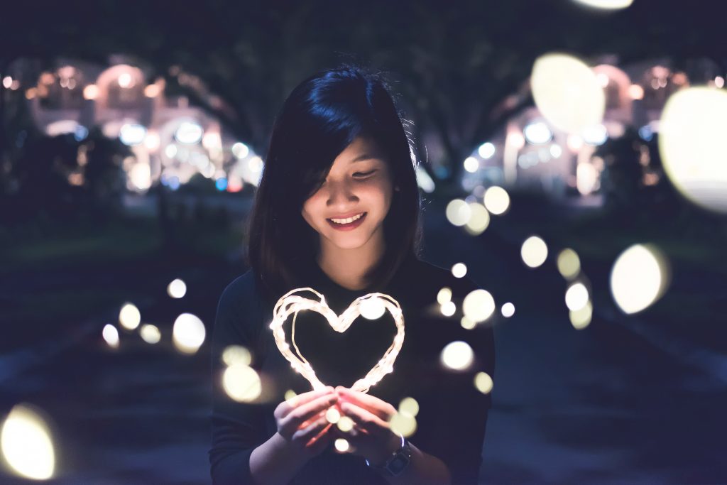 Auf diesem Bild ist eine junge Frau zu sehen, die ein leuchtendes Herz in der Hand hält, welches sie lächelnd anschaut.