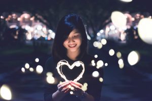 Auf diesem Bild ist eine junge Frau zu sehen, die ein leuchtendes Herz in der Hand hält, welches sie lächelnd anschaut.