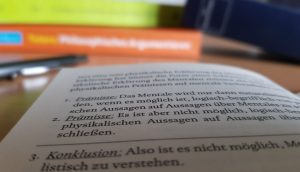 Zu sehen ist ein aufgeschlagenes Buch mit Fachliteratur. Im Hintergrund kann man weitere Bücher erahnen.