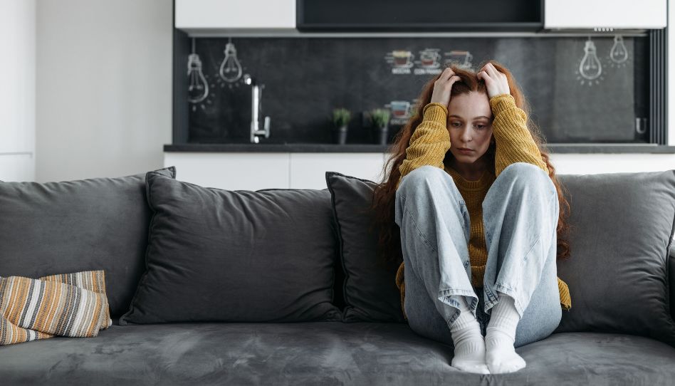Eine junge Frau sitzt mit angewinkelten Beinen auf dem Sofa, die Hände auf dem Kopf. Sie sieht traurig aus