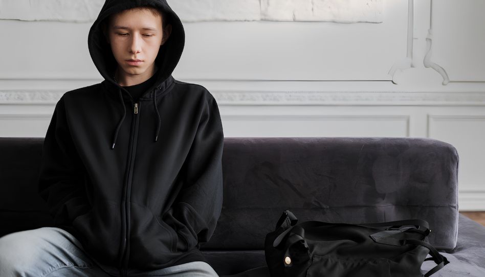 Ein junger Mann mit dunklem Kapuzenpullover sitzt auf einem Sofa. Er schaut traurig nach unten.