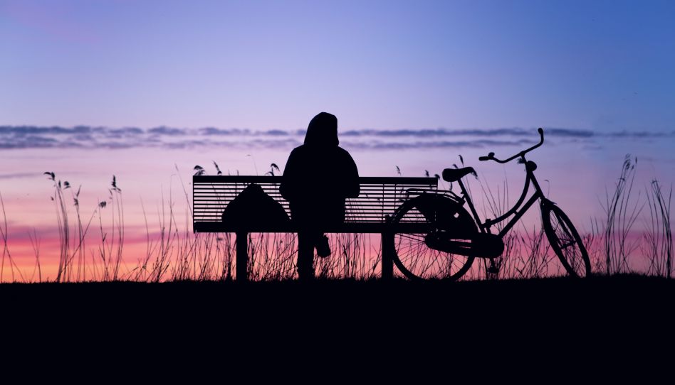 Eine Silhouette einer Person von hinten, die auf einer Bank sitzt. Neben der Bank steht ein Fahrrad. Im Hintergrund sieht man den Himmel eines Sonnenuntergangs.