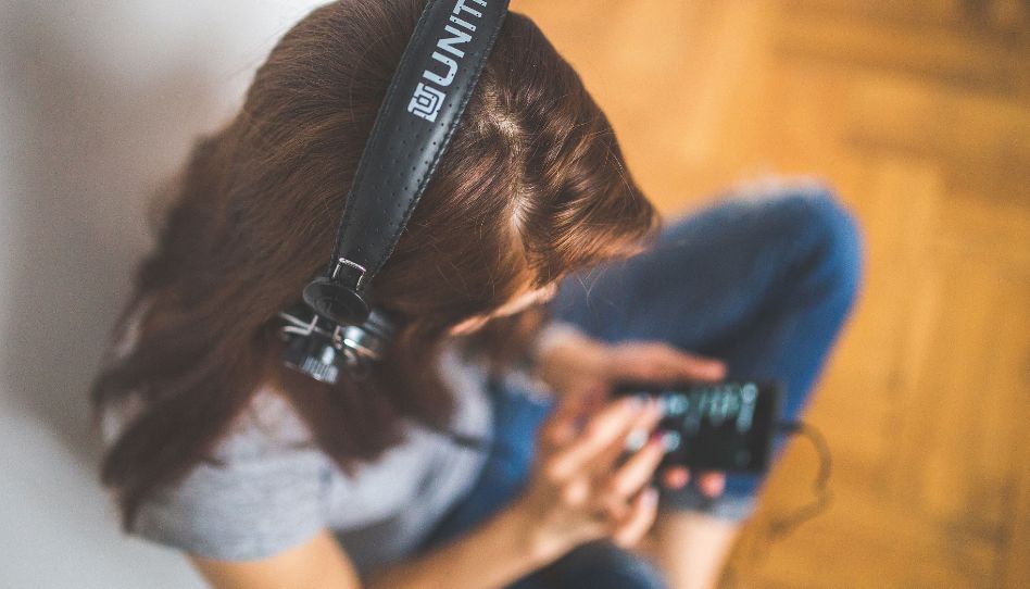 Eine junge Frau sitzt im Schneidersitz. Sie hat Kopfhörer auf und schaut auf ihr Handy.