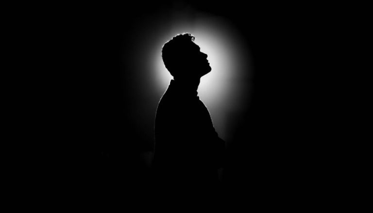 Man sieht lediglich den oberen Teil der Silhouette eines Mannes. Im Hintergrund ist eine Lichtquelle, sonst ist es komplett dunkel. Der Mann schaut nach oben.