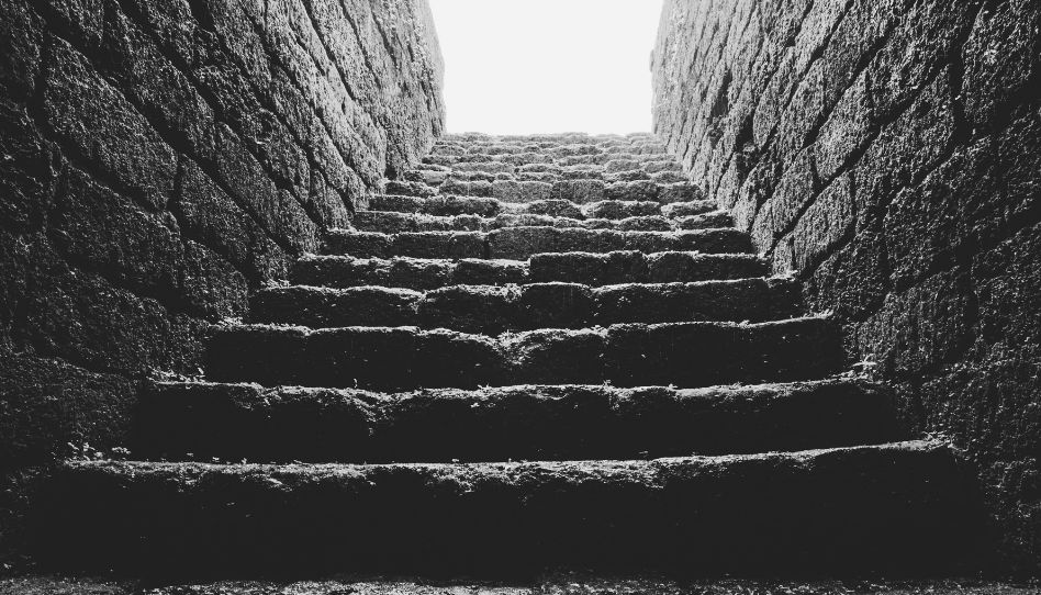 Eine Treppe, die nach oben führt. Links und rechts sind Wände. Oben geht man in Richtung des Lichts. Das Foto ist schwarz-weiß.