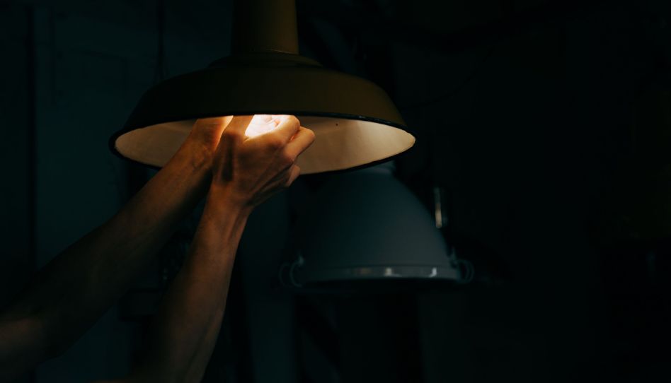 Zwei Hände an der Birne einer Deckenlampe. Die Glühbirne leuchtet – sonst ist es dunkel.