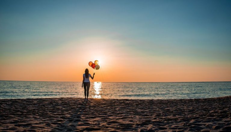 Eine Frau steht, mit dem Rücken zur Kamera, an einem Strand vorm Meer. Sie hält ein paar Luftballons in der Hand.