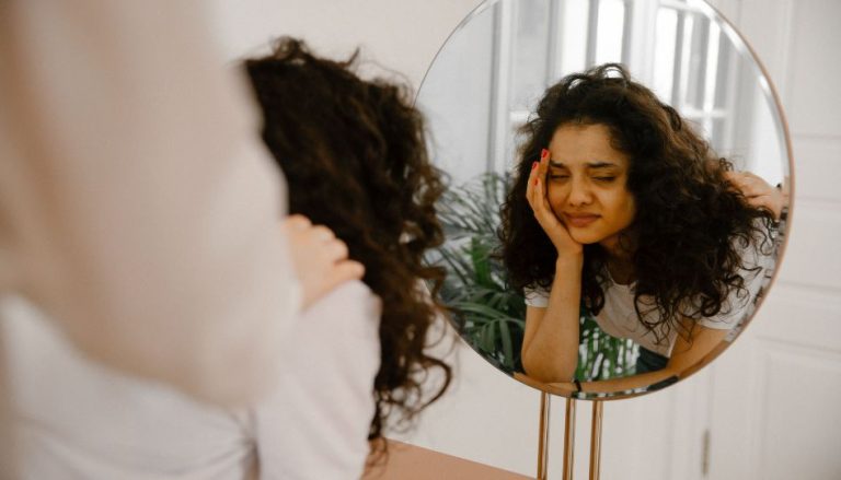 Man sieht das Spiegelbild einer jungen Frau. Sie hat den Kopf auf ihrer rechten Hnd abgestützt und die Augen verschlossen. Sie sieht traurig aus. Jemand hat die Hand auf ihrer Schulter.