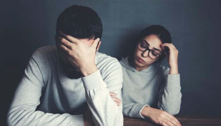 Ein junger Mann ist auf einem Tisch nach vorne gebeugt und hält sich die linke Hand vor das Gesicht. Eine junge Frau, die neben ihm sitzt, lehnt an der Wand, hat die Augen verschlossen und ihren Kopf leicht auf die Linke Hand gestützt. Sie hat einen traurigen Gesichtsausdruck.