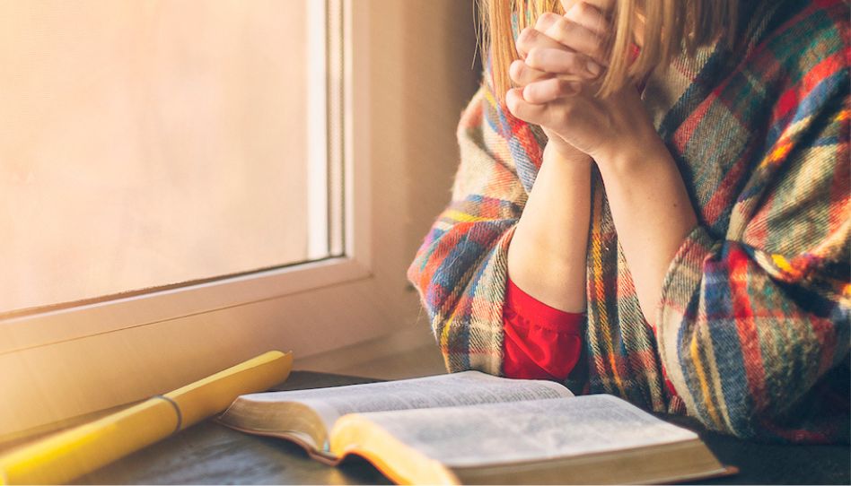 Eine Frau hat die Hände zum Beten gefaltet. Vor ihr auf dem Tisch liegt eine geöffnete Bibel.